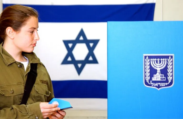 Elecciones en Israel: una soldado deposita su voto junto a una cabina de votación con el emblema del Estado y la bandera israelí