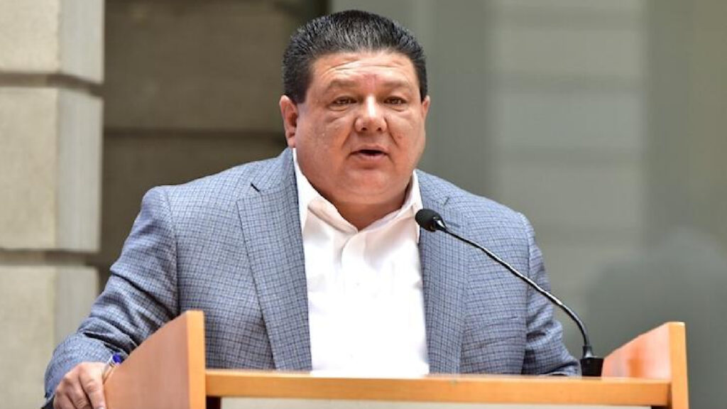 Exfiscal de Chihuahua por traición a la patria: César Jáuregui Moreno habla tras un atril con micrófono, con saco gris y camisa blanca