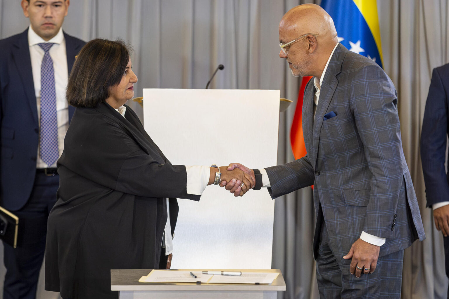 Dinorah Figuera y Jorge Rodríguez se estrechan la mano en la mesa del diálogo político en Venezuela, con la bandera venezolana detrás