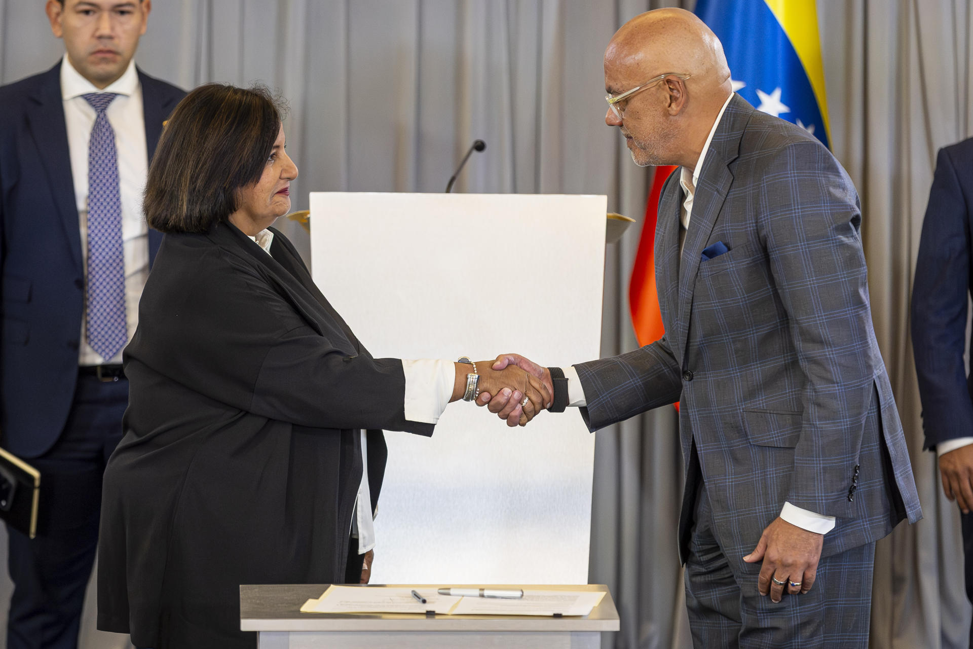 Dinorah Figuera y Jorge Rodríguez se estrechan la mano en la mesa del diálogo político en Venezuela, con la bandera venezolana detrás
