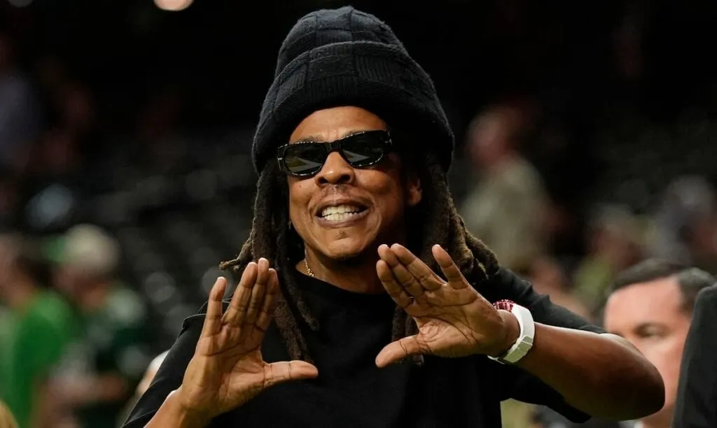 Jay-Z en el Super Bowl: el rapero, con gorro negro y gafas de sol, hace su característica seña con las manos en un evento deportivo