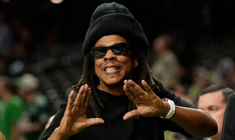 Jay-Z en el Super Bowl: el rapero, con gorro negro y gafas de sol, hace su característica seña con las manos en un evento deportivo