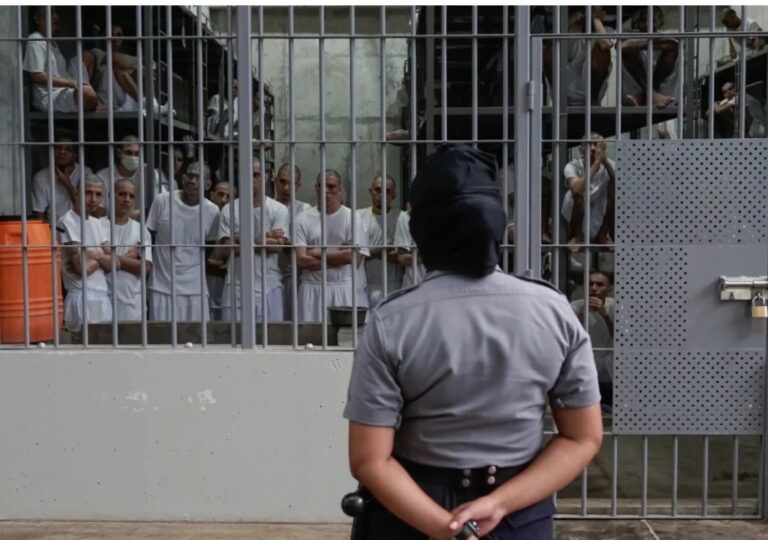 Pandilleros condenados en El Salvador: reos de blanco tras las rejas de una prisión salvadoreña vigilados por una agente penitenciaria