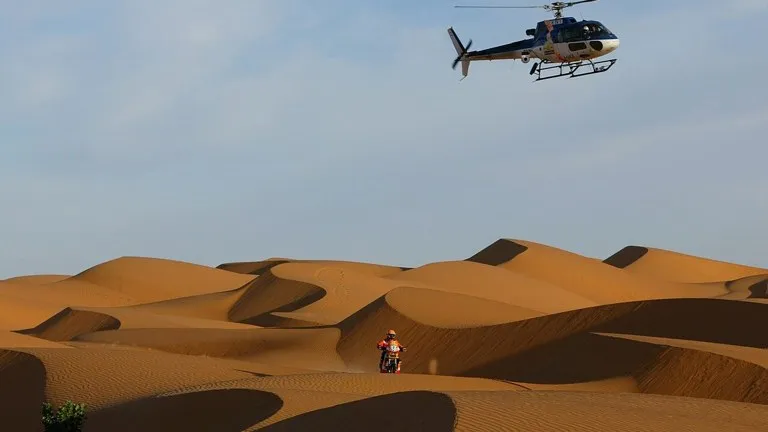 Un piloto de motos cruza las dunas del desierto seguido por el helicóptero de la organización, la imagen clásica que el Rally Dakar 2027 recuperará en Marruecos
