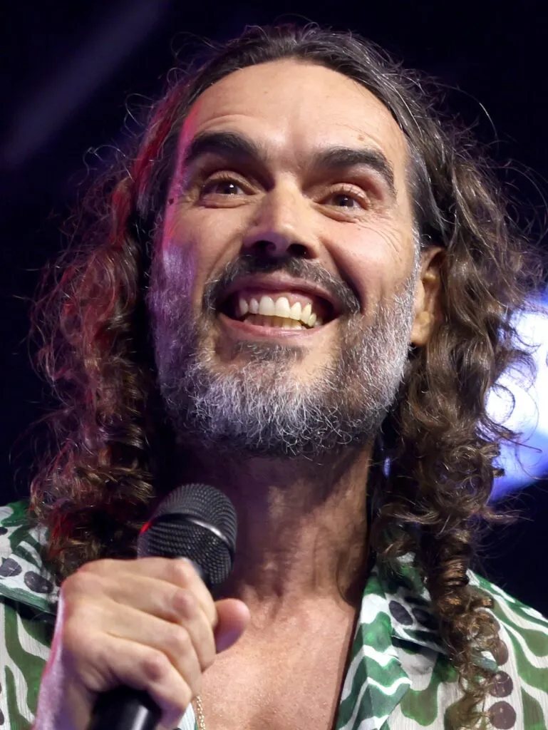 Russell Brand, declarado no culpable de un cargo de agresión indecente: el comediante británico sostiene un micrófono en un escenario en 2025